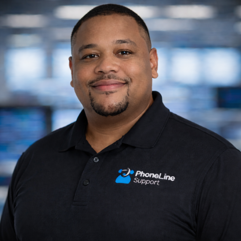 A man in a black polo shirt with a PhoneLine Support logo smiles at the camera, standing indoors with a blurred background of blue and gray tones.